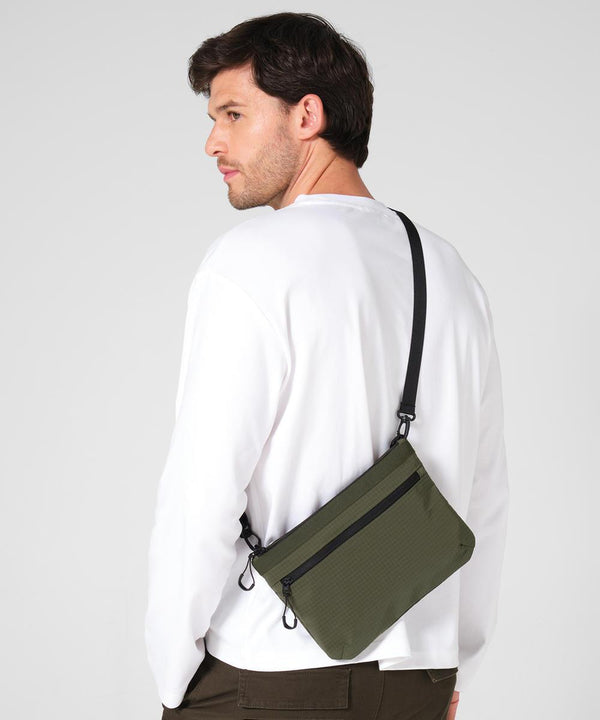 A man with short brown hair wears a white long-sleeve shirt, dark trousers, and carries the Quadra UrbanShift sacoche—an olive green crossbody bag with a black zip and strap. The background is plain light grey.