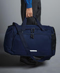 A person holds a large navy blue Quadra QD449 duffel bag with double zippers and shoulder strap. The bag has a side mesh pocket and small front pocket. The person wears dark pants, a blue sweater, and black shoes.