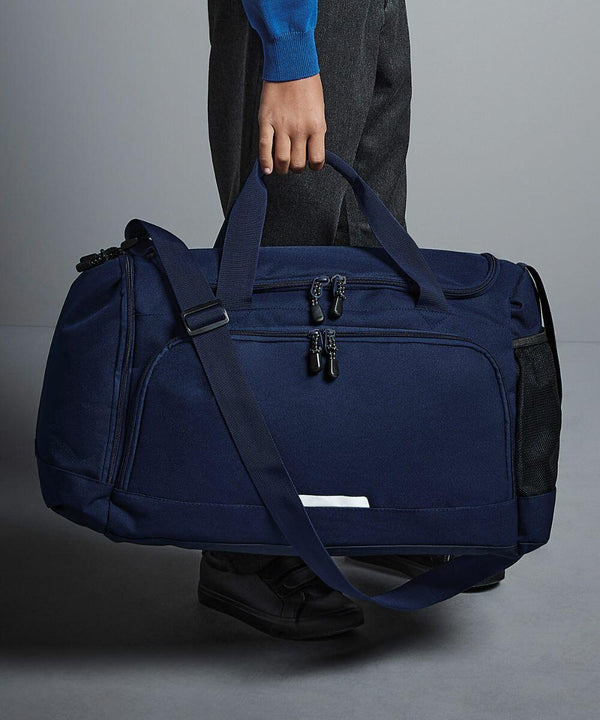 A person holds a large navy blue Quadra QD449 duffel bag with double zippers and shoulder strap. The bag has a side mesh pocket and small front pocket. The person wears dark pants, a blue sweater, and black shoes.