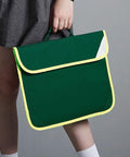 A person holds a green Quadra QD452 satchel-style school bag with yellow trim. The photo is cropped to show only the lower part of the person's body and the bag.