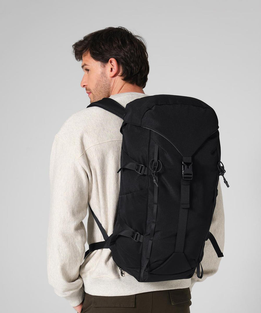 A person in a light beige jacket and dark trousers is seen from the back carrying the Quadra Active Outdoor 30 litre rucksack, which is black with multiple straps and buckles, against a plain light grey background.