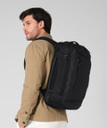 A man in a tan jacket and white trousers carries the Quadra Zrich travel rucksack over his shoulder, standing against a light grey background.