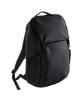 The Bags black rucksack features a top handle, front zip pocket, and two side pockets. Photographed on a white background with visible shoulder straps and a closed main compartment.