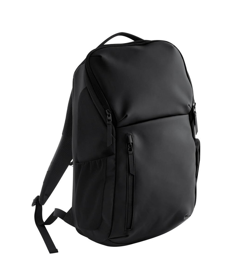 The Bags black rucksack features a top handle, front zip pocket, and two side pockets. Photographed on a white background with visible shoulder straps and a closed main compartment.
