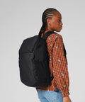 A person with plaited hair, dressed in a brown checked shirt with blue flower patterns and blue jeans, carries a Quadra Zrich toploader rucksack. They stand sideways against a plain light background.