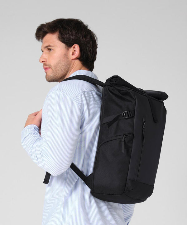 A man with short brown hair in a light blue striped shirt is shown in profile, carrying the Quadra Zrich roll-top rucksack over his shoulder against a plain grey background.