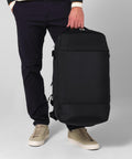 A person in dark trousers and beige trainers holds a Quadra Zrich 45 litre hybrid holdall by the top handle, standing against a plain light background.