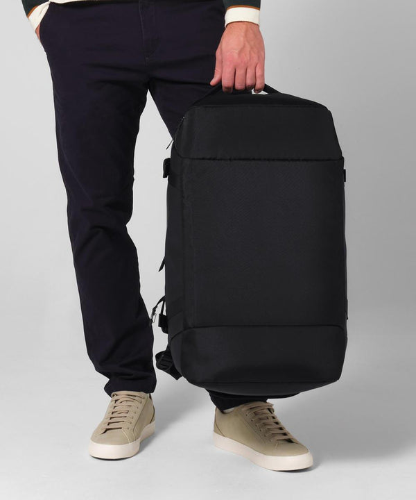A person in dark trousers and beige trainers holds a Quadra Zrich 45 litre hybrid holdall by the top handle, standing against a plain light background.