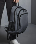 A person holding a grey Quadra QD550 backpack with multiple compartments and black zippers, dressed in dark pants and white sneakers, standing on a grey floor against a grey background.