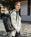 Young man with blond hair wearing a light-colored jacket and carrying a large black Quadra QD552 backpack, standing outdoors on a paved area near a building.
