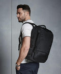 A man with short dark hair and a beard is wearing a white shirt and carrying a large Quadra QD565 backpack on his right shoulder. He is facing to the left against a plain gray background.