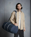 A man with long hair and a beard stands against a gray wall, wearing a beige button-up shirt over a white and black striped tee, and carrying a large blue Quadra QD613 over his shoulder.