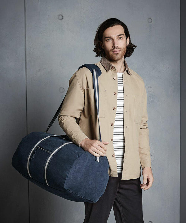 A man with long hair and a beard stands against a gray wall, wearing a beige button-up shirt over a white and black striped tee, and carrying a large blue Quadra QD613 over his shoulder.