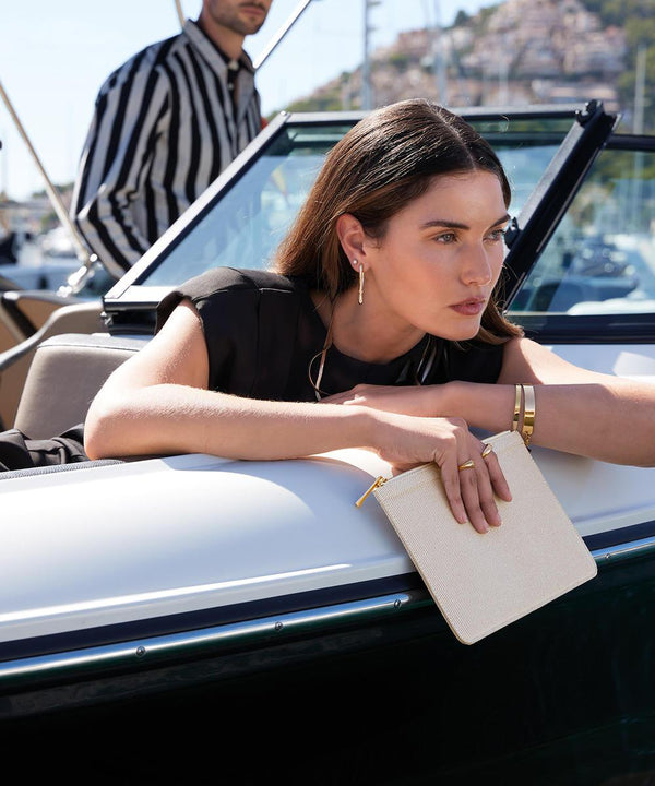 A woman with long brown hair, dressed in black and wearing gold jewellery, leans on a boat holding the Quadra Puerto pouch. In the background, a man wears a black and white striped shirt.