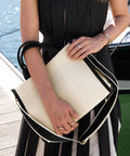 Wearing a black dress with white pleats, a person stands on a quay holding the Quadra Puerto medium tote in beige and black, accessorised with multiple rings and bracelets. A boat and water appear in the background.