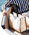 A person in a black-and-white striped shirt and white shorts sits on a beige and black boat seat with the Quadra Puerto large tote, a cream bag with black handles and trim, resting on their lap.
