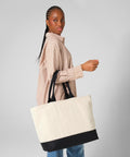 A woman in a loose, striped beige shirt and black trousers stands sideways, holding the Quadra Puerto weekender—a large beige tote bag with black handles and a black bottom—against a plain light grey background.