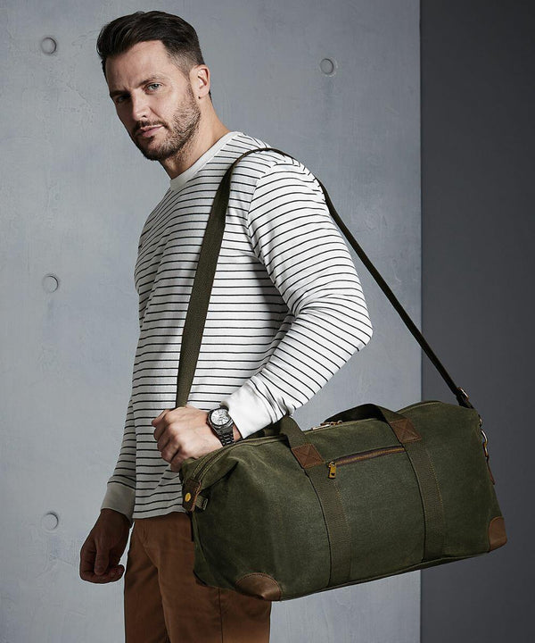 A man wearing a striped long-sleeve shirt is holding a green Quadra QD650 duffel bag on his shoulder and looking towards the camera against a plain background.