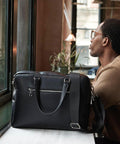 A man with short hair and glasses sits by a window, looking outside. In front of him on the table is a Quadra Tailored Luxe PU briefcase featuring a shoulder strap and silver zips.