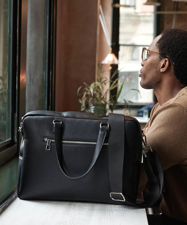 A man with short hair and glasses sits by a window, looking outside. In front of him on the table is a Quadra Tailored Luxe PU briefcase featuring a shoulder strap and silver zips.
