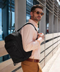 A person wearing sunglasses, a light peach shirt, and tan trousers stands outside a modern building, carrying the Quadra Airside underseat rucksack over one shoulder and looking off to the side.