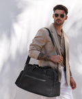 A man stands against a white wall carrying the Quadra Airside weekender holdall over his shoulder, wearing sunglasses, a beige blazer, taupe shirt, and white trousers.
