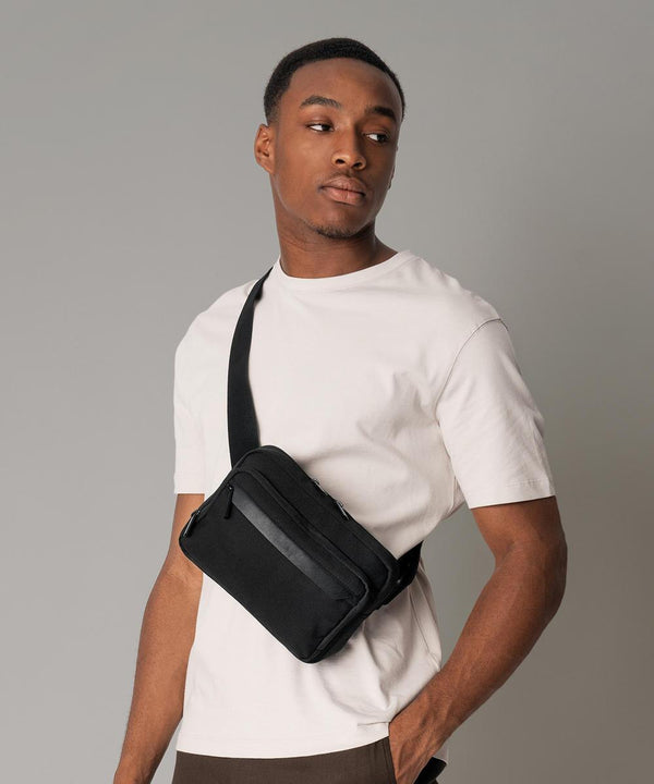 A person wearing a white short-sleeve t-shirt stands against a plain grey background, looking to the side with one hand resting on the Quadra Airside cross body bag in black.