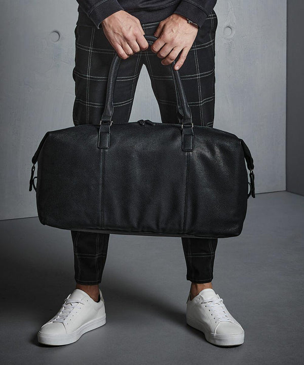 A person holding a black leather Quadra QD878, wearing black checkered pants and white sneakers, standing on a gray surface.
