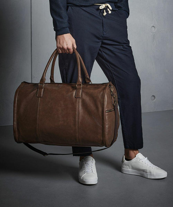 A person in dark blue pants and white sneakers holds a large brown Quadra QD880 leather duffel bag.