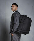 A person wearing a dark jacket and gray pants stands against a plain background, carrying a large black Quadra QD902 with straps over one shoulder.