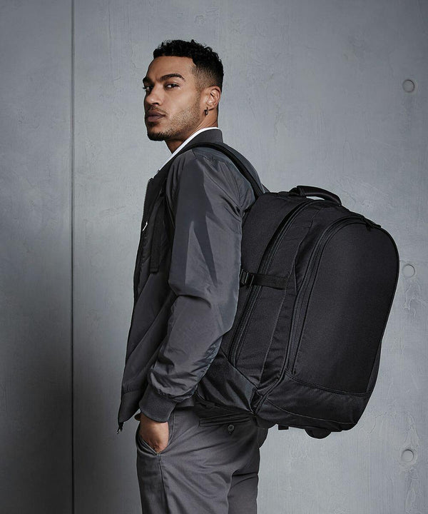 A person wearing a dark jacket and gray pants stands against a plain background, carrying a large black Quadra QD902 with straps over one shoulder.