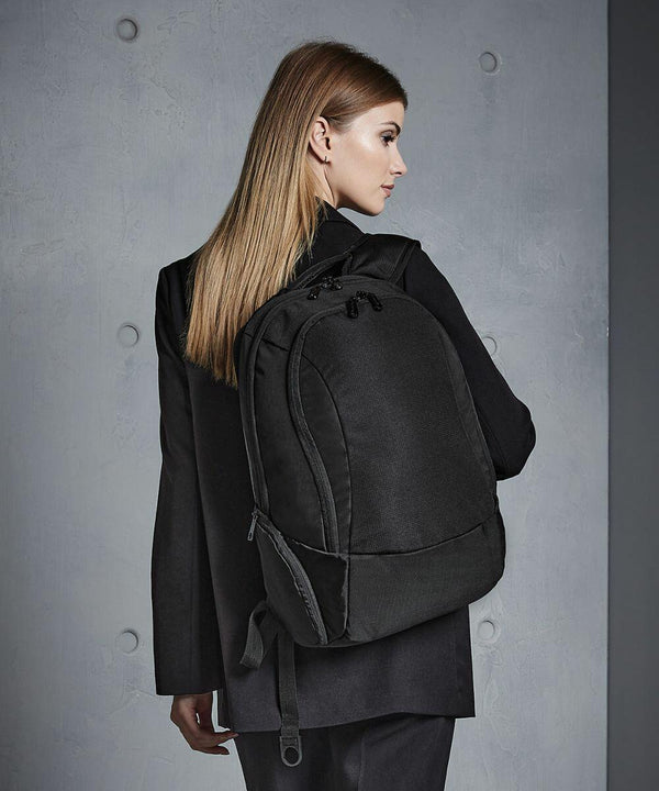 A woman with straight, light brown hair wears a black blazer and carries a large black Quadra QD906 backpack, standing against a gray background.