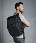 A man with a beard and short hair is standing sideways, arms crossed, wearing a striped short-sleeve shirt and a large black Quadra QD910 backpack against a gray textured background.