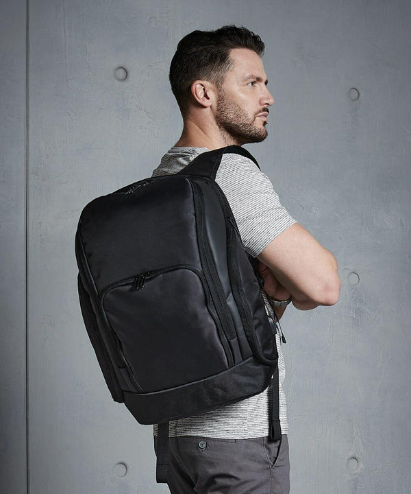 A man with a beard and short hair is standing sideways, arms crossed, wearing a striped short-sleeve shirt and a large black Quadra QD910 backpack against a gray textured background.