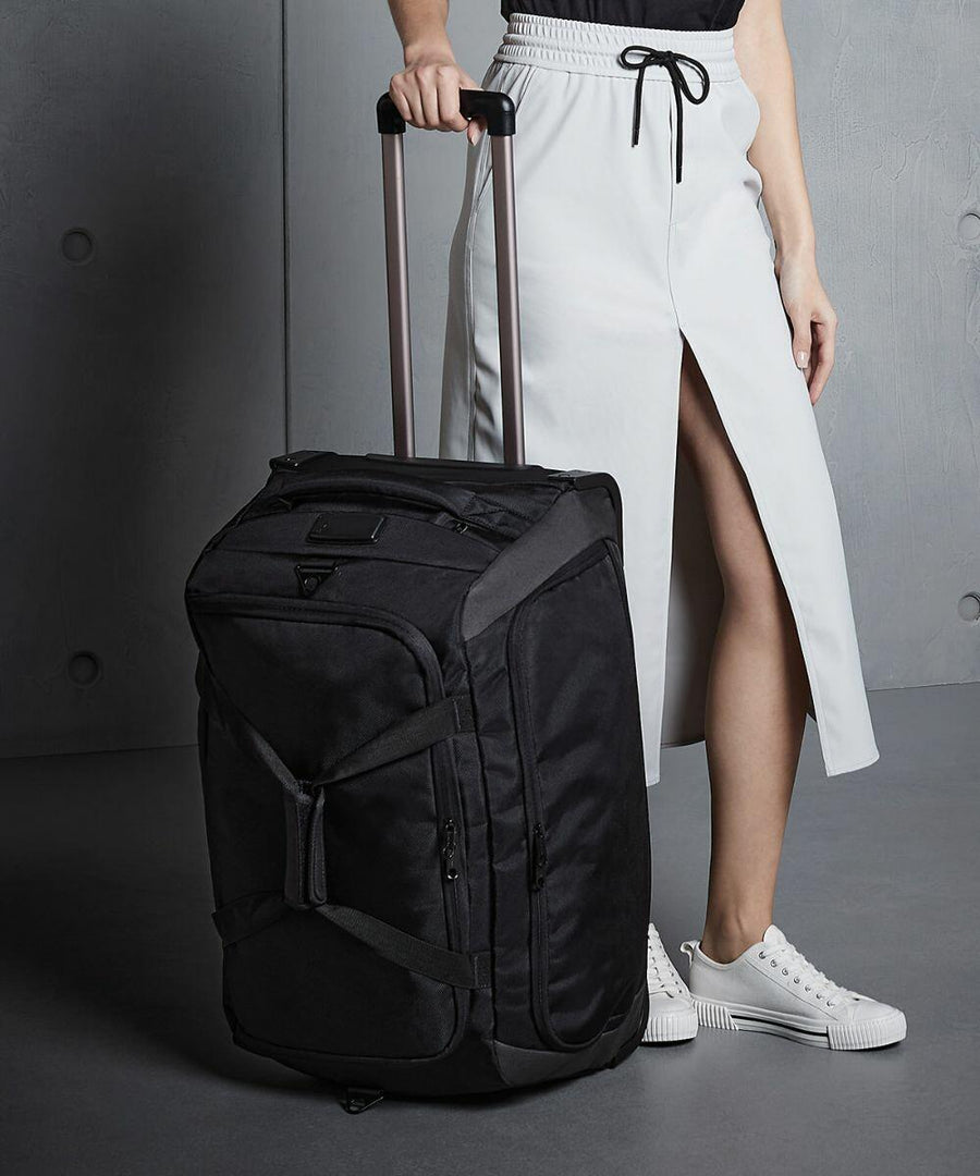 A person in a white skirt and white sneakers stands beside a black Quadra QD970 wheeled suitcase, holding its extended handle.
