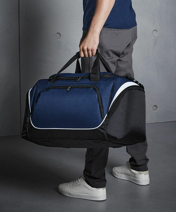 A person in a blue shirt and gray pants is holding a large, black and blue Quadra QS270 duffle bag. White sneakers are visible on the concrete floor.