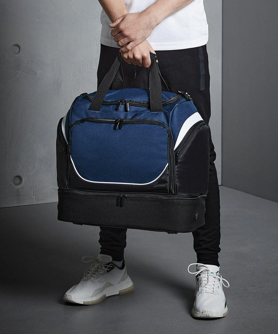 Person holding a blue and black Quadra QS285 gym bag with multiple compartments, wearing a white shirt, black pants, and white sneakers.