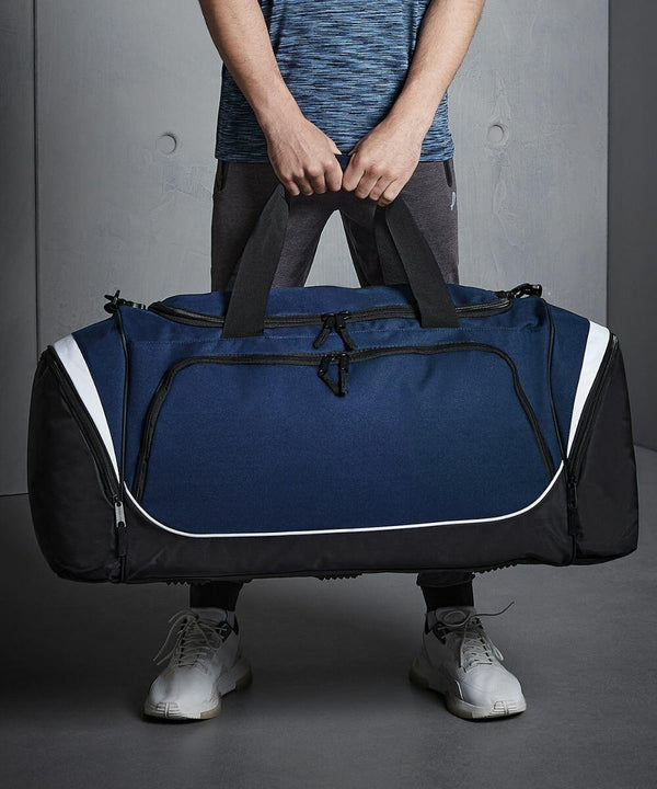 Person holding a large blue and black Quadra QS288 duffel bag with both hands. The person is wearing a blue shirt, grey pants, and white sneakers.