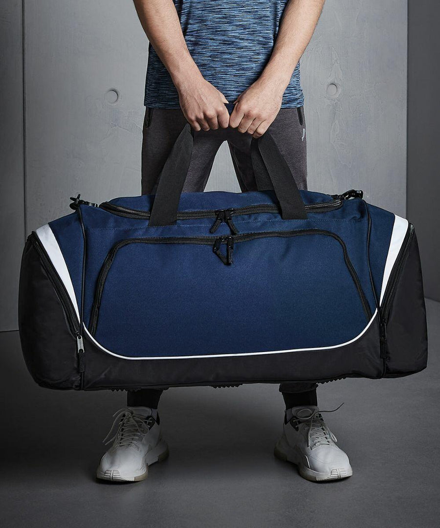 Person holding a large blue and black Quadra QS288 duffel bag with both hands. The person is wearing a blue shirt, grey pants, and white sneakers.