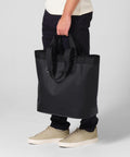 A person wearing black trousers, a light shirt, and beige trainers holds the Quadra Waterproof 35 litre tote bag by its handles. Only their lower half is visible against a plain light background.