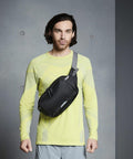 Person with medium-length hair wearing a yellow long-sleeve shirt and grey pants, posing against a plain grey background with a Quadra QX515 black crossbody bag worn across the chest.