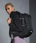 Person with short hair wearing a black hoodie and purple pants, carrying a large black Quadra QX520 backpack over one shoulder, stands against a grey wall.