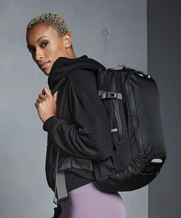 Person with short hair wearing a black hoodie and purple pants, carrying a large black Quadra QX520 backpack over one shoulder, stands against a grey wall.
