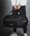 Person in a white jacket and black pants holding a large black Quadra QX560 duffel bag with both hands. The background is a plain, grey wall.