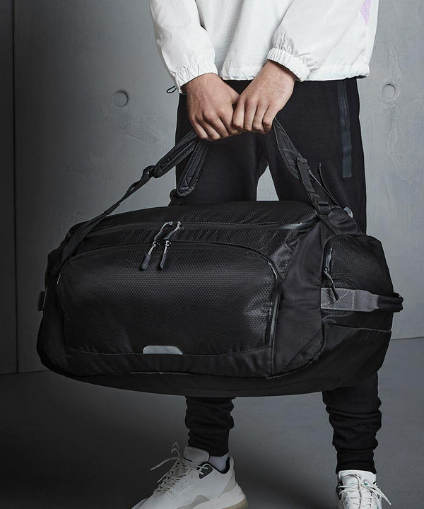 Person in a white jacket and black pants holding a large black Quadra QX560 duffel bag with both hands. The background is a plain, grey wall.
