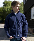 A young man in a navy blue Result R114J zip-up jacket stands outside, looking to the side with his hands in his pockets. A building and trees are visible in the background.
