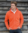 Person in a Result R114X orange zip-up jacket and jeans stands with hands in pockets against a weathered wooden wall.
