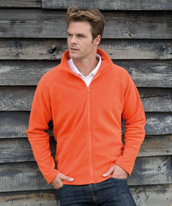 Person in a Result R114X orange zip-up jacket and jeans stands with hands in pockets against a weathered wooden wall.