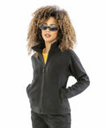 A person with curly hair is wearing sunglasses, a black Result R115F zip-up jacket, a yellow shirt, and dark pants, standing with hands in pockets against a white background.