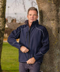 A man wearing a navy blue Result R118A jacket stands outdoors, leaning against a tree. He is zipping up his jacket, with trees and a grassy area in the background.